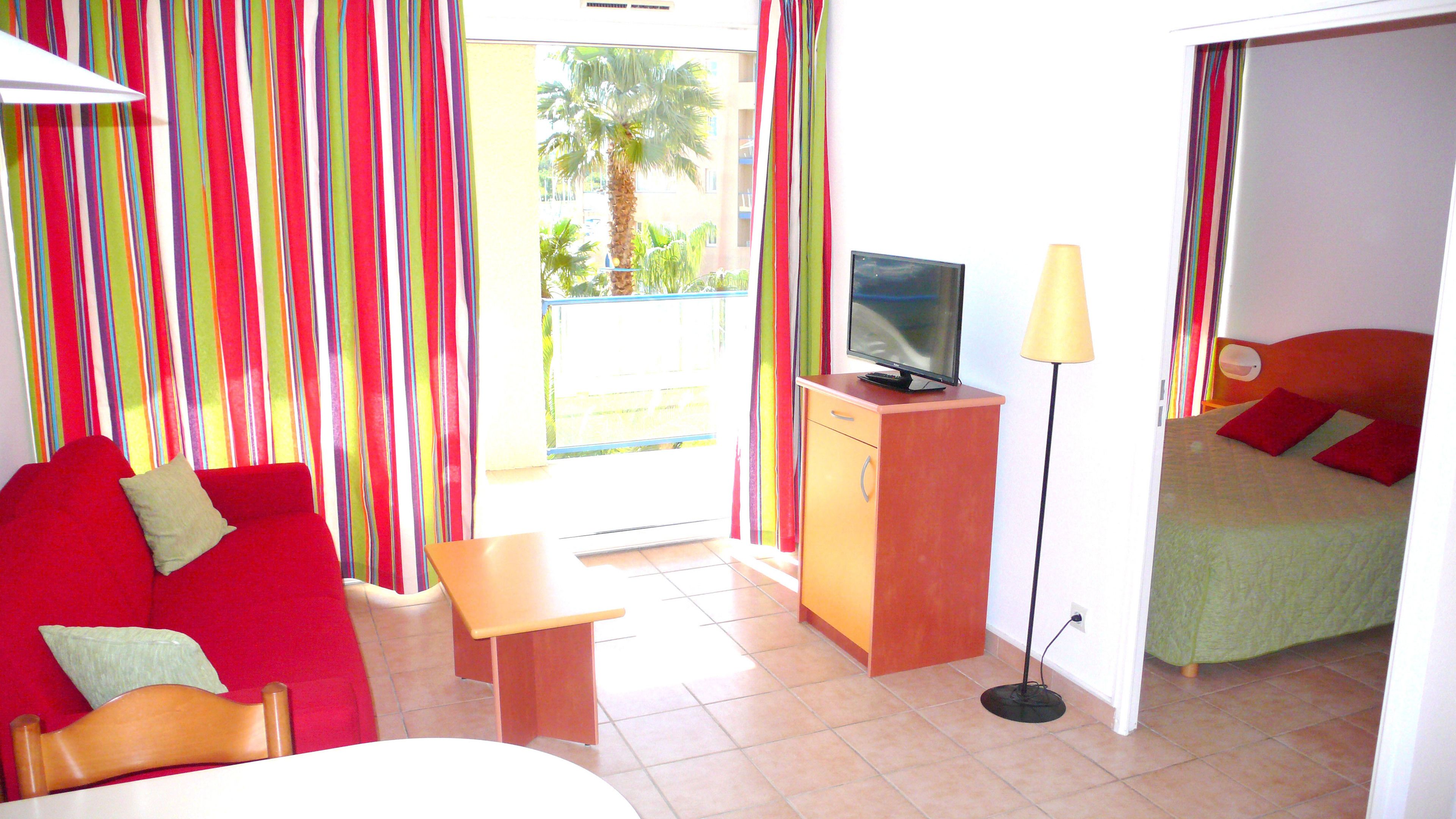 A hotel room with a bed, a TV on a wooden stand, a small table, a lamp, and a colorful curtain.