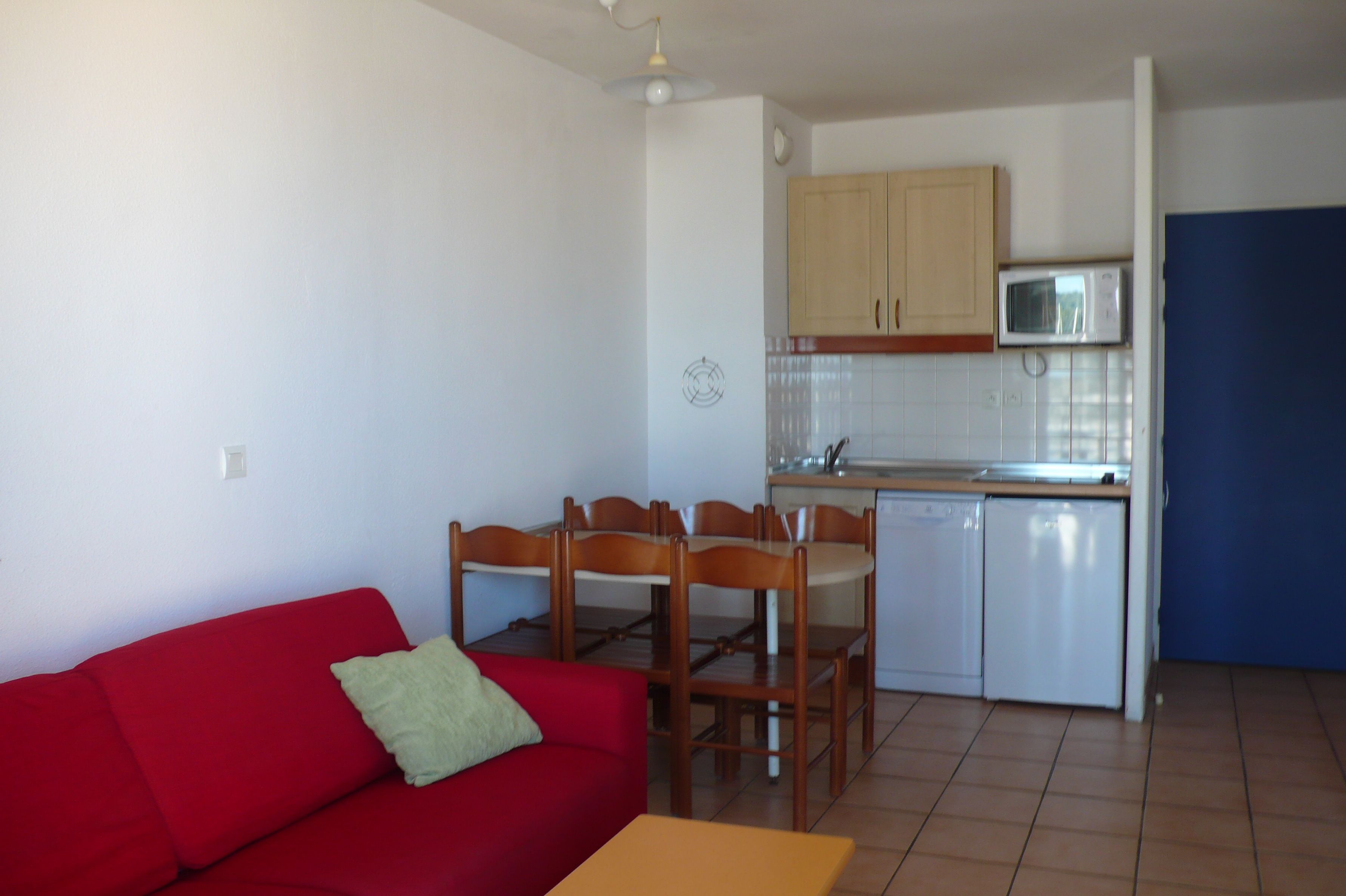 A compact living space with a red sofa, wooden dining table, and chairs. A kitchen area with a microwave and cabinets is visible.
