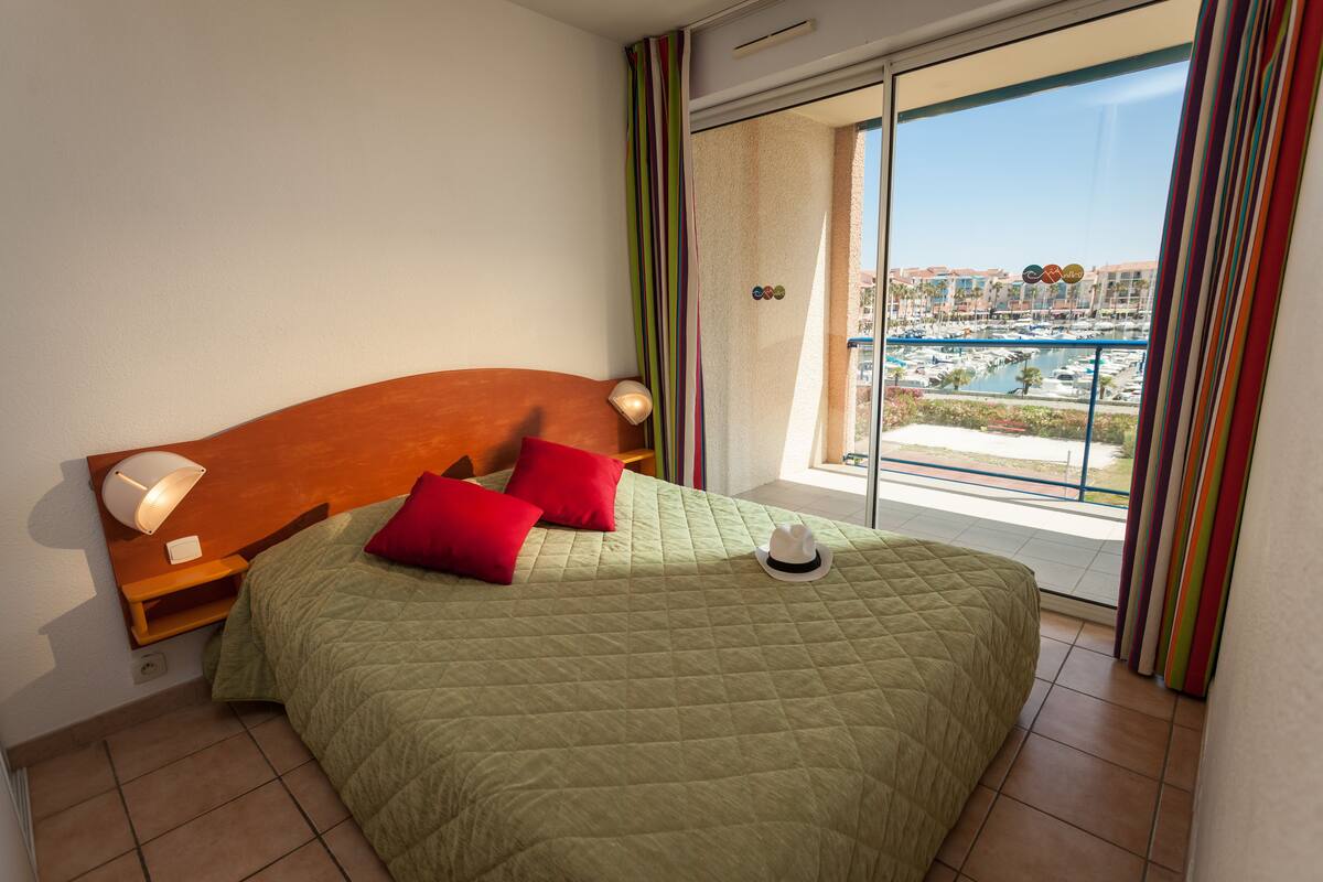 Ein Schlafzimmer mit einem großen Bett, einem Nachttisch, einer Lampe und Blick auf eine Marina vom Balkon aus.