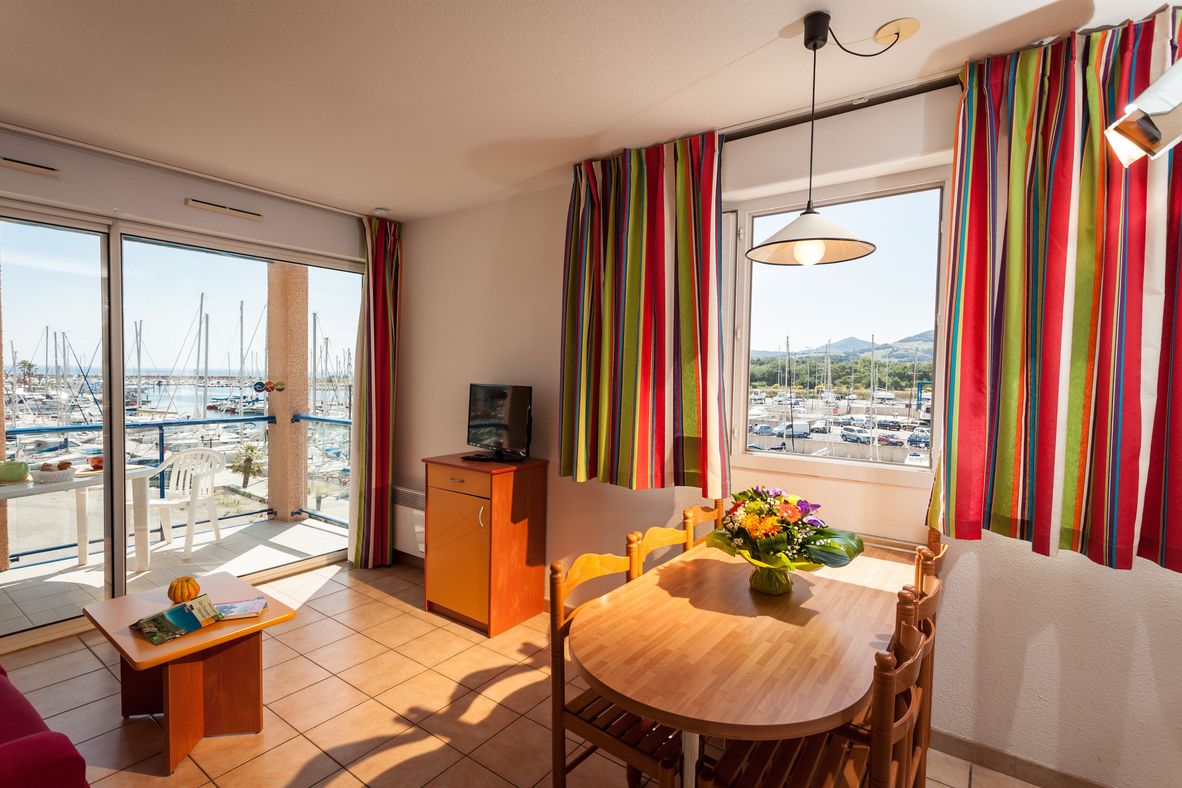 Ein modernes Wohnzimmer mit Essbereich, großen Fenstern und bunten Vorhängen, einem Fernseher auf einer Kommode und Blick auf eine Marina.