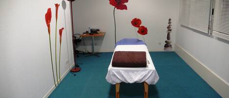 Una habitación con una cama, un escritorio y una silla. Hay flores rojas en la pared y una ventana con persianas.