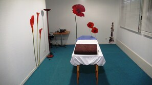 A room with a bed, a desk, and a chair. There are red flowers on the wall and a window with blinds.