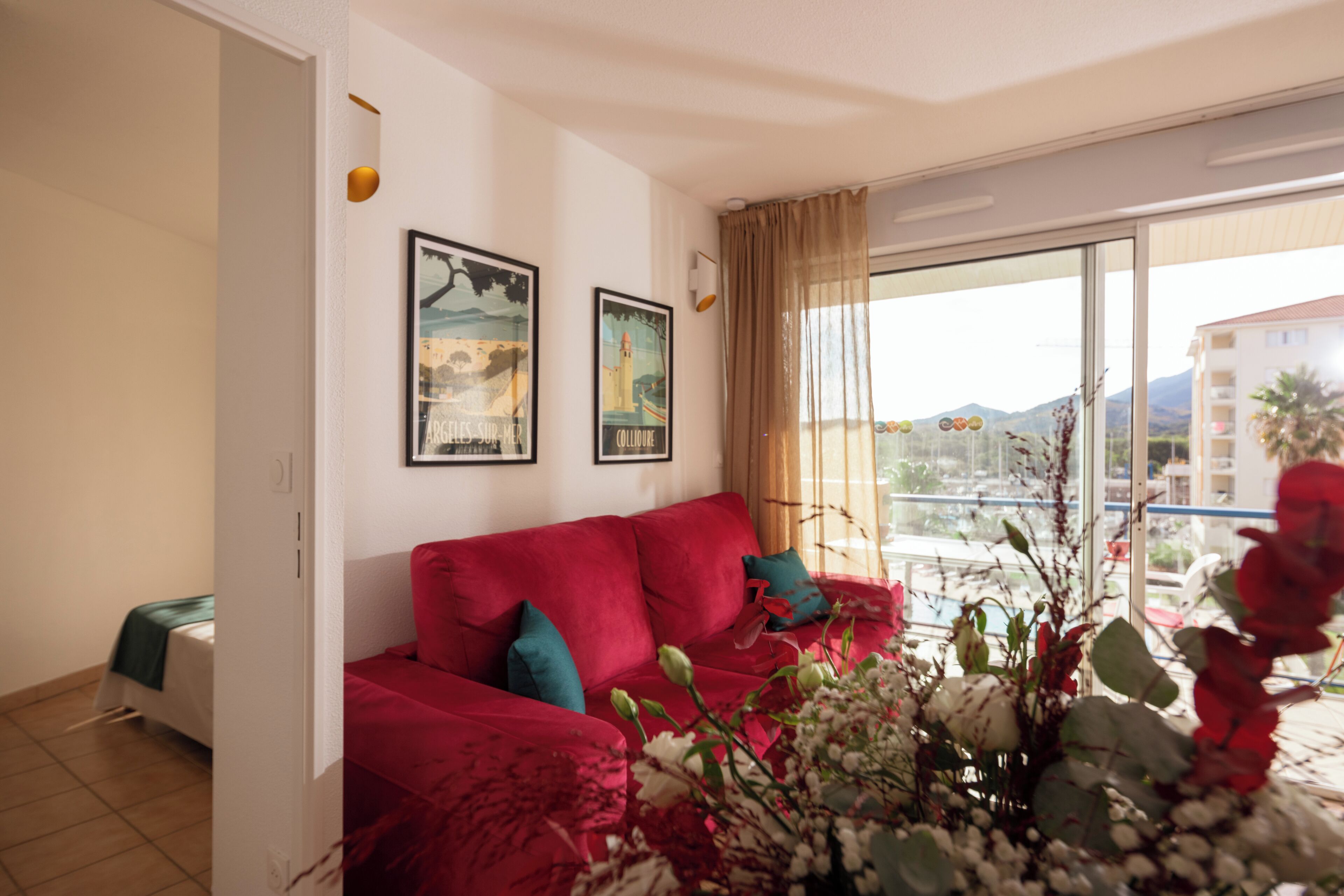 A living room with a red sofa, two framed pictures on the wall, a large window with a view of buildings and mountains, and a balcony with plants.