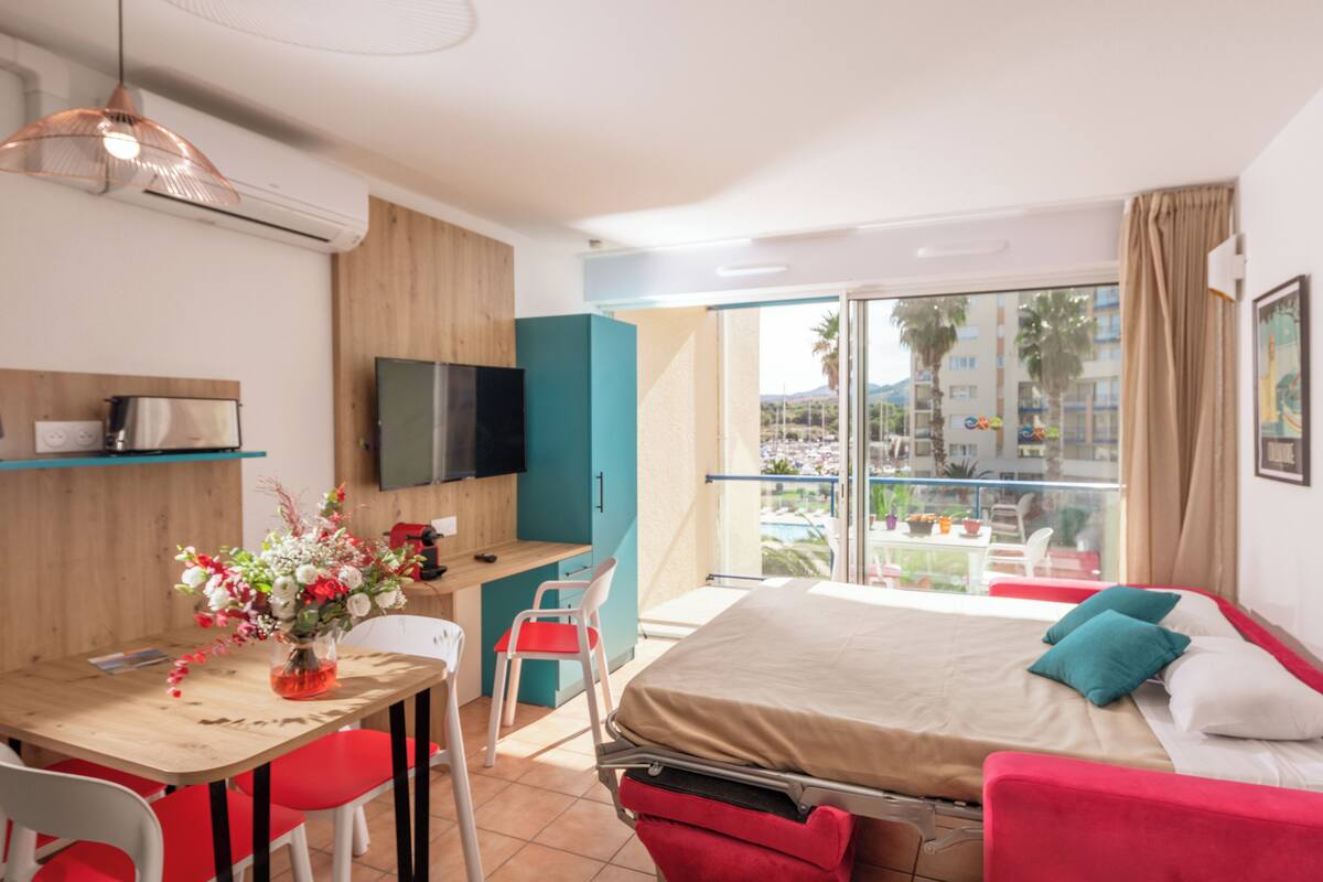 Ein modernes Hotelzimmer mit einem Bett, einem Schreibtisch mit Stuhl, einem Fernseher und einem Balkon mit Blick auf Palmen und Gebäude.