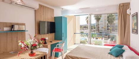 A modern hotel room with a bed, a desk with a chair, a TV, and a balcony with a view of palm trees and buildings.