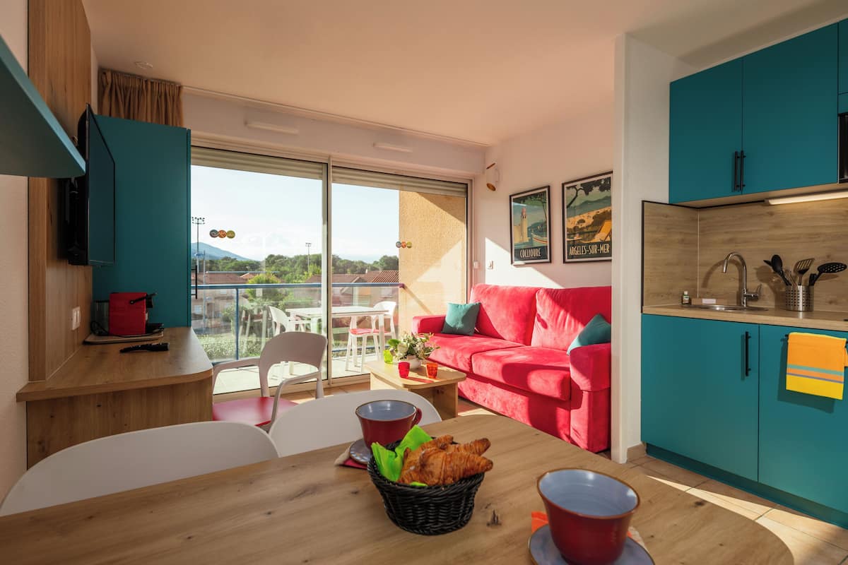 Ein moderner Wohnraum mit einem roten Sofa, türkisfarbenen Schränken, einem Holzesstisch mit Croissants und einem Balkon mit Blick.