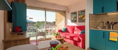 Un espacio habitable moderno con un sofá rojo, armarios color turquesa, una mesa de comedor de madera con cruasanes y un balcón con vistas.