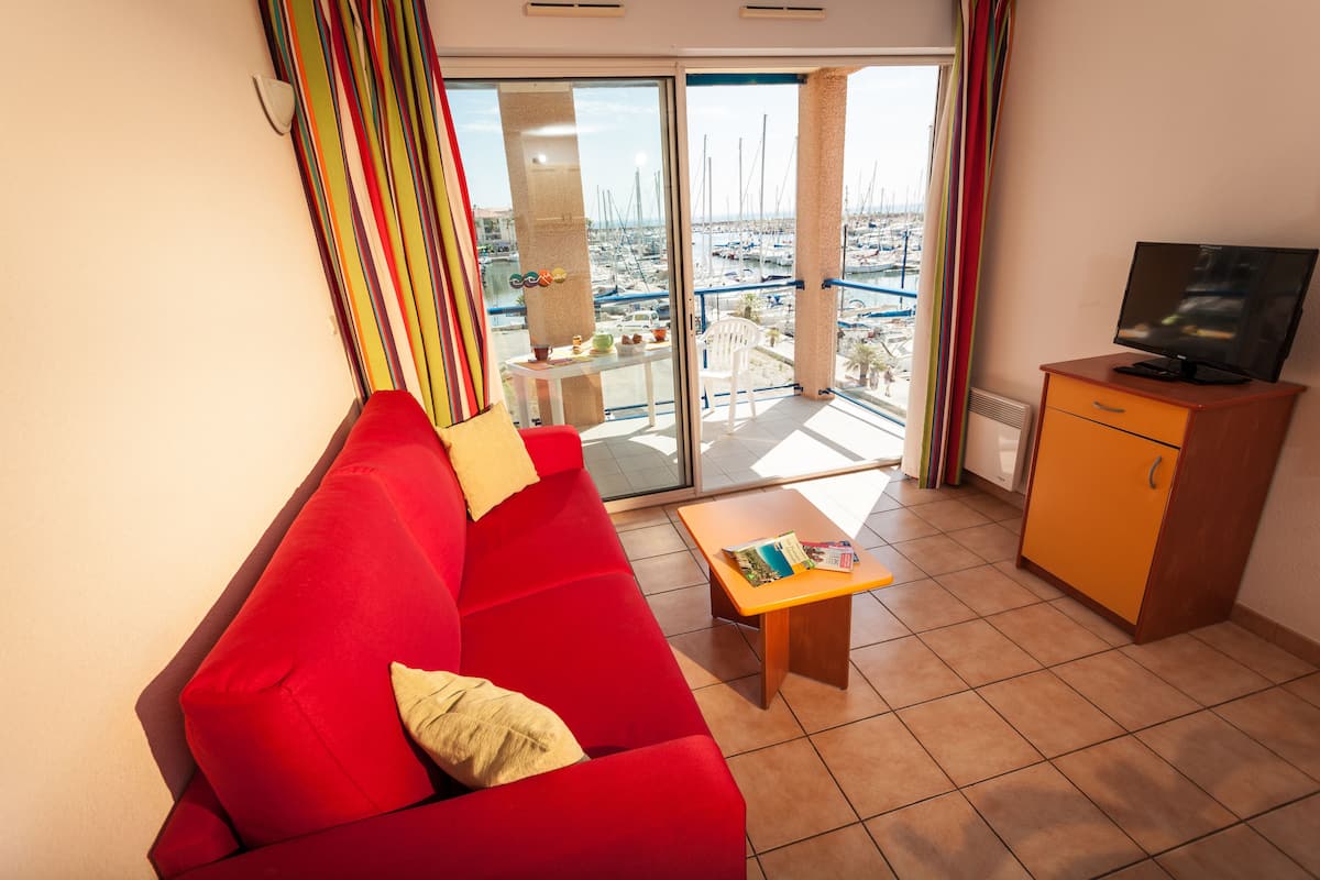 Ein rotes Sofa, ein kleiner Tisch mit Zeitschriften, ein Fernseher auf einer Holzkommode und ein Balkon mit Blick auf Boote und Gebäude.