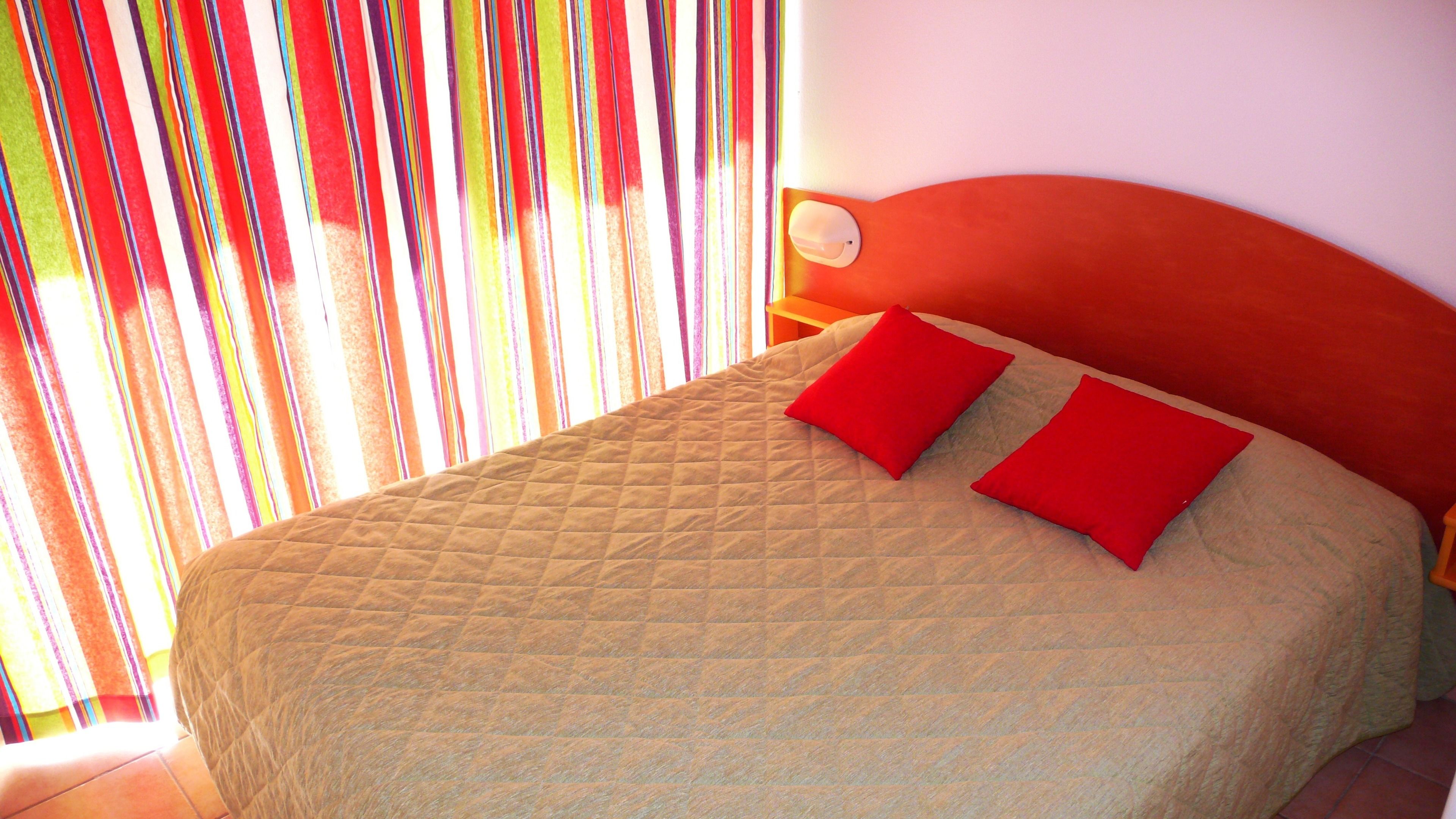 Una cama con cubrecolcha acolchada y dos almohadas rojas, cabecera de madera y ventana con persianas verticales.