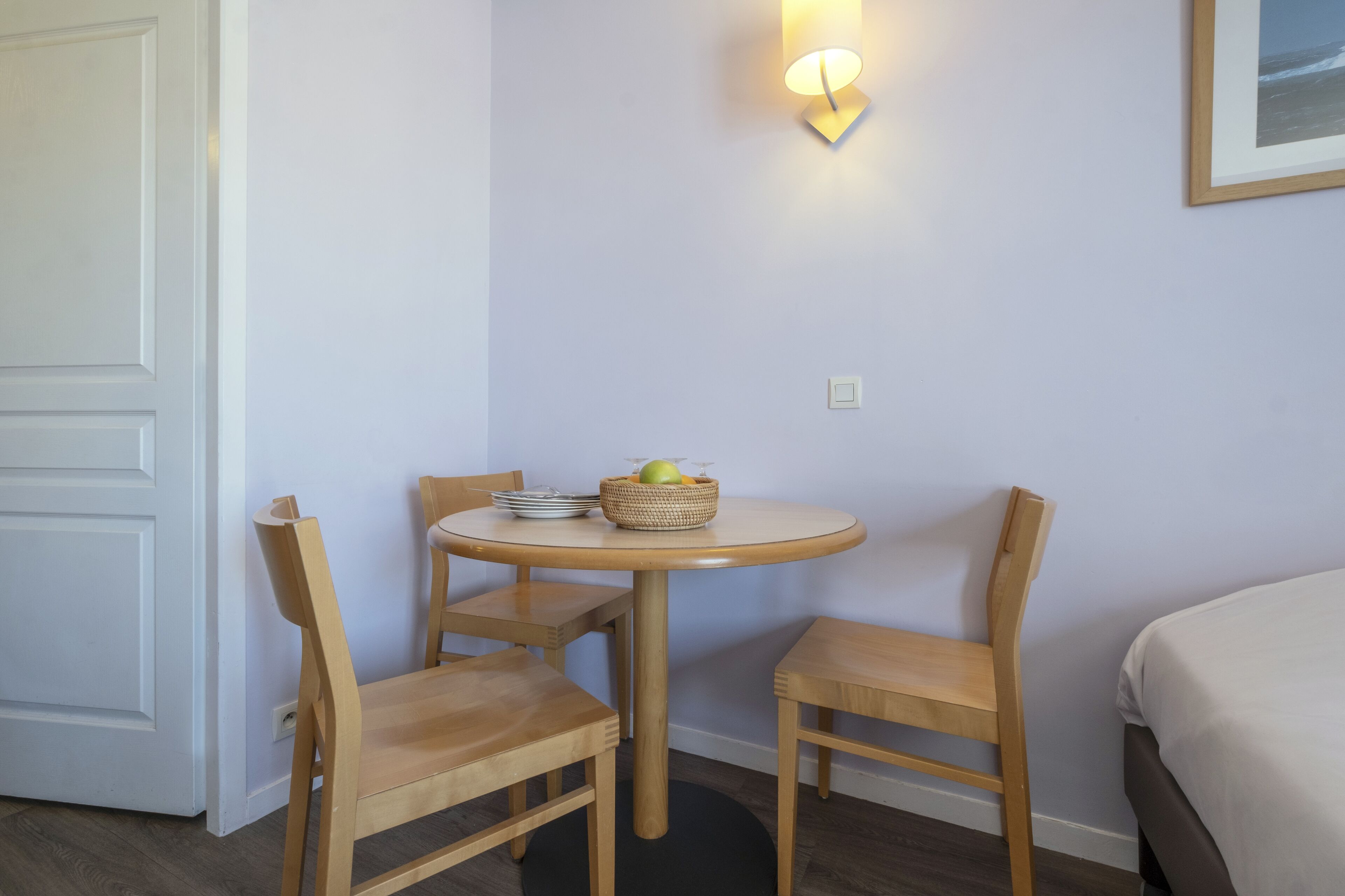Et lille værelse med et rundt bord, to træstole, en vægmonteret lampe og en seng.