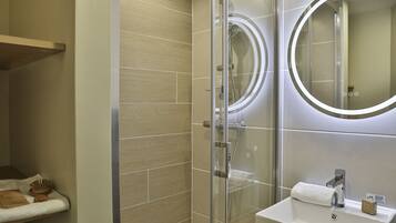 A modern bathroom with a glass-enclosed shower, a round mirror with built-in lighting, and a white sink with a soap dispenser.
