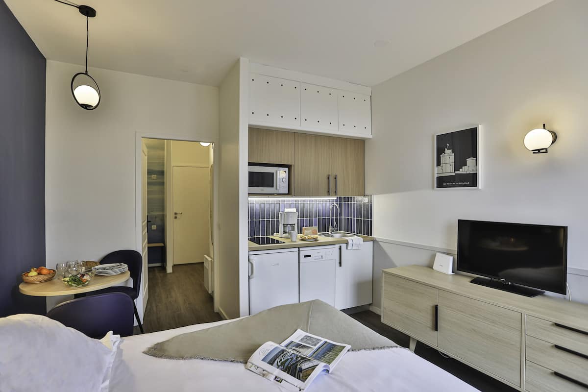 A modern hotel room with a bed, a dining area, a kitchenette, and a TV.