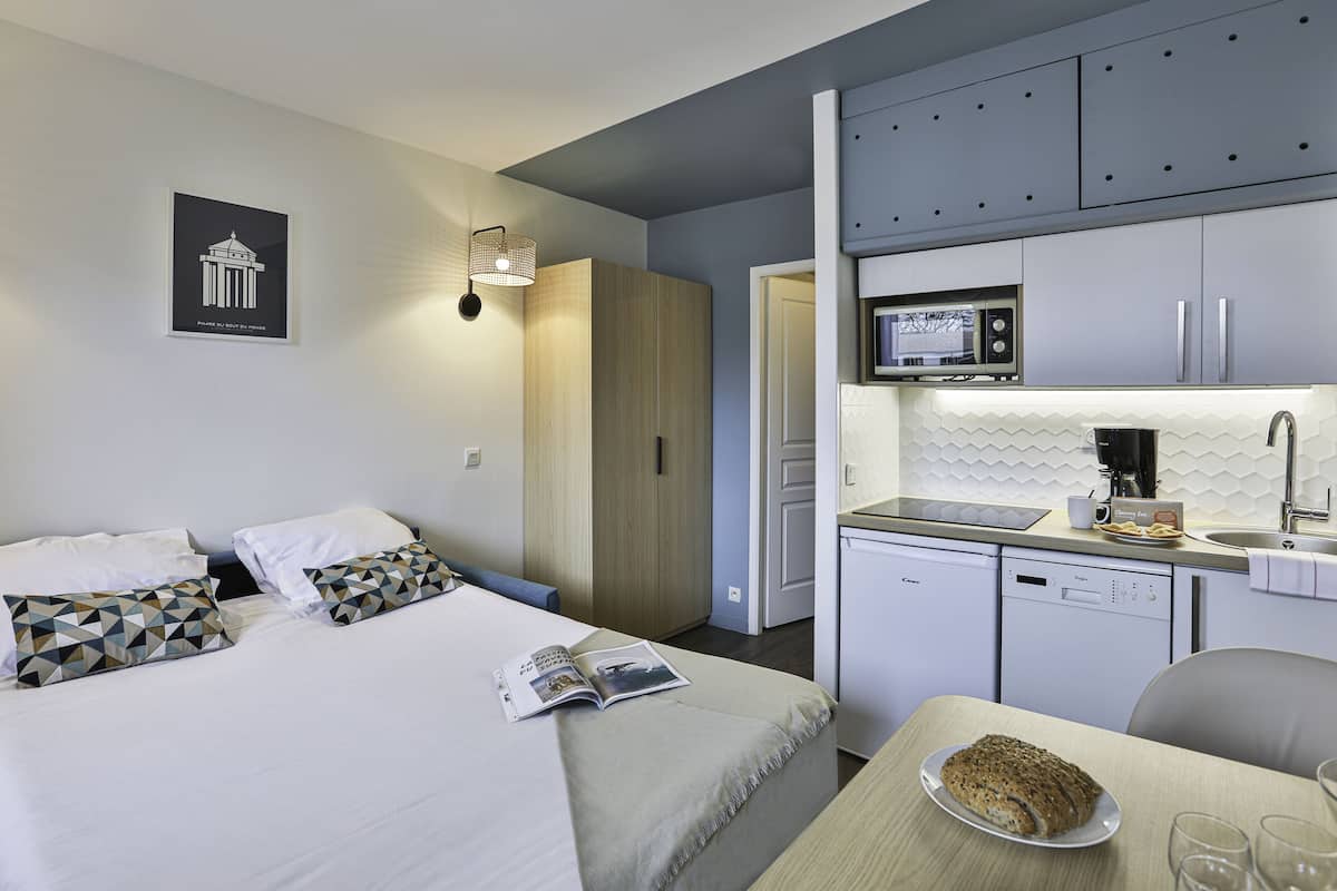 A modern hotel room with a bed, a small kitchenette, and a dining area.