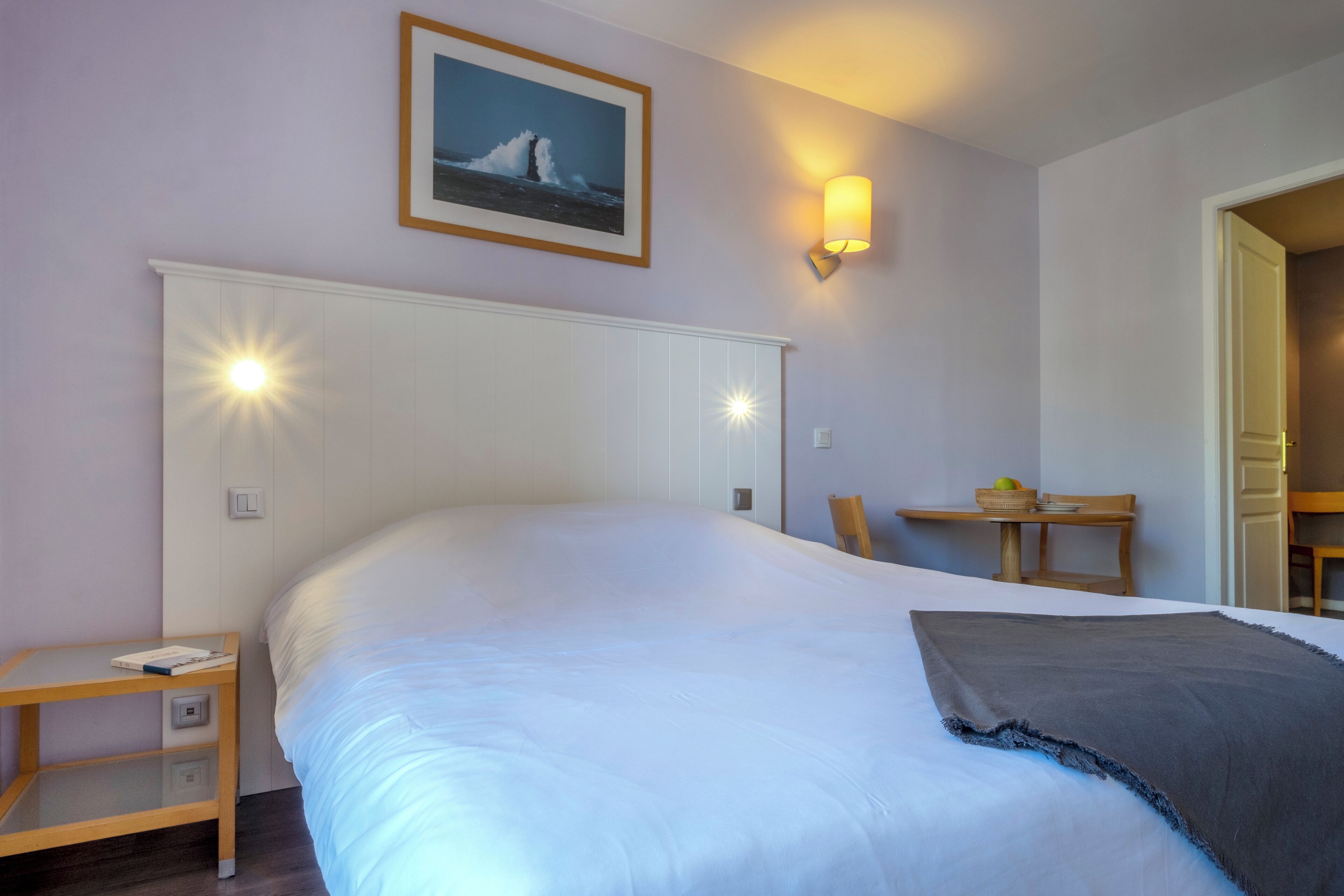 Une chambre d’hôtel avec un grand lit, une table de chevet, une chaise, une petite table avec un panier et une lampe fixée au mur.