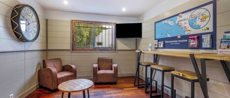 A room with a wooden floor, two leather chairs, a round table, a map on the wall, and a TV mounted above a wooden bar area.