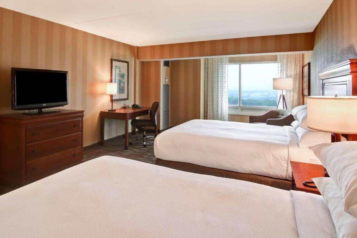 A hotel room with two beds, a desk, a television, and a view of the city.