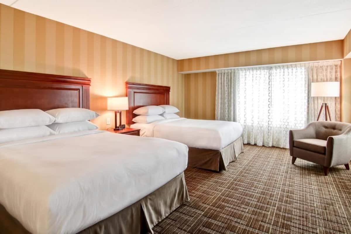 A hotel room with two beds, a chair, a lamp, and a window with curtains.