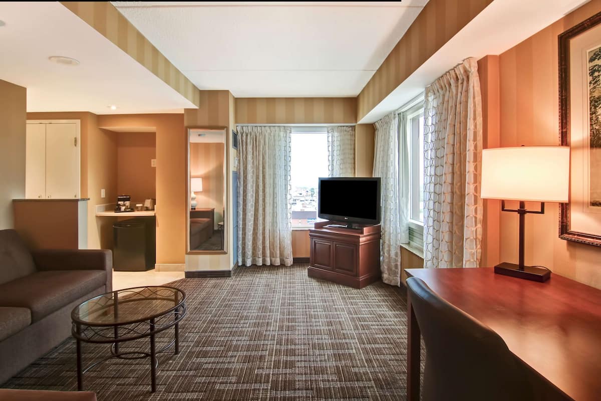 A hotel room with a sofa, a small table, a TV, a lamp, and a view of the outside through a window with curtains.