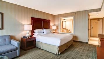 A hotel room with a large bed, two bedside tables with lamps, a gray armchair, and a mirror on the wall.