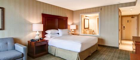 A hotel room with a large bed, two bedside tables with lamps, a gray armchair, and a mirror on the wall.