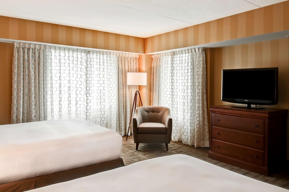 A hotel room with two beds, a chair, a TV on a dresser, and curtains with a geometric pattern.