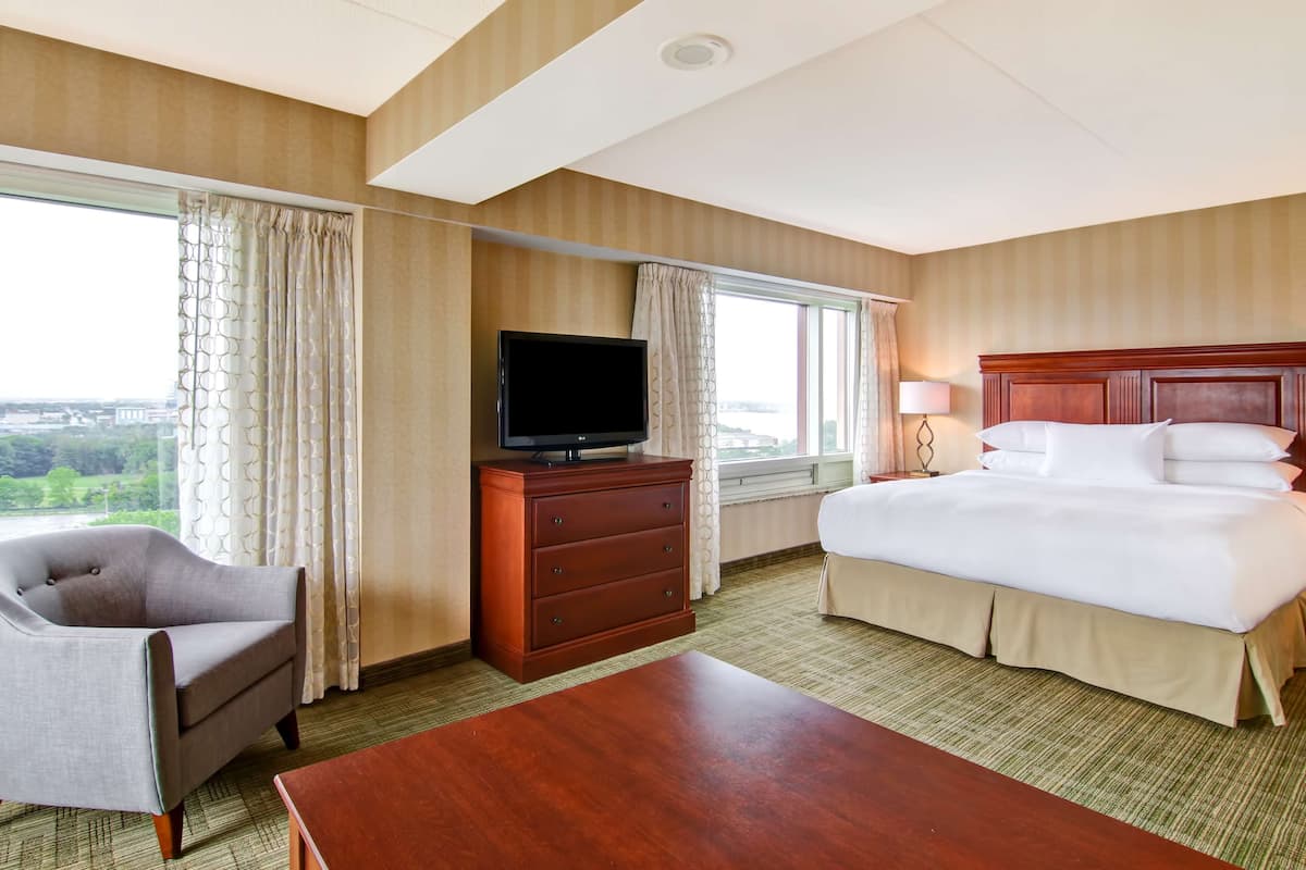 A hotel room with a bed, a TV on a dresser, a chair, and a large window offering a city view.