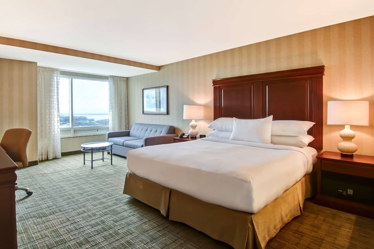 A hotel room with a large bed, a sofa, a small table, and a desk. There are two lamps on the desk and a window with curtains.