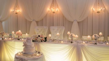 A wedding reception setup with a tiered cake, floral centerpieces, and draped white curtains.
