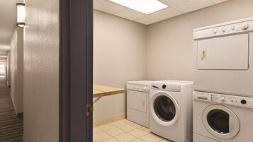 A laundry room with two washing machines, a wooden shelf, and a tiled floor.