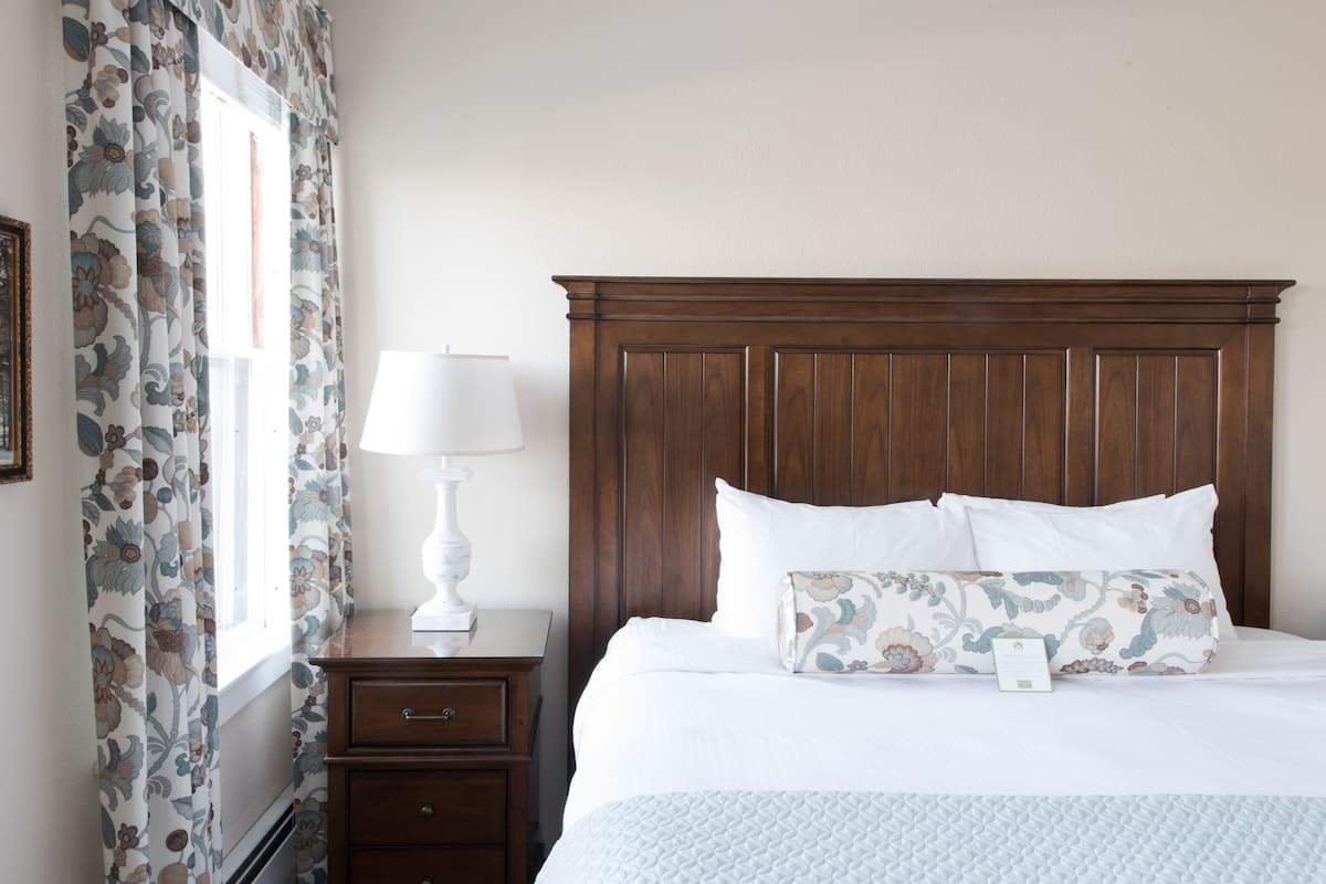 A neatly made bed with a wooden headboard, a bedside table with a lamp, and a floral-patterned curtain.