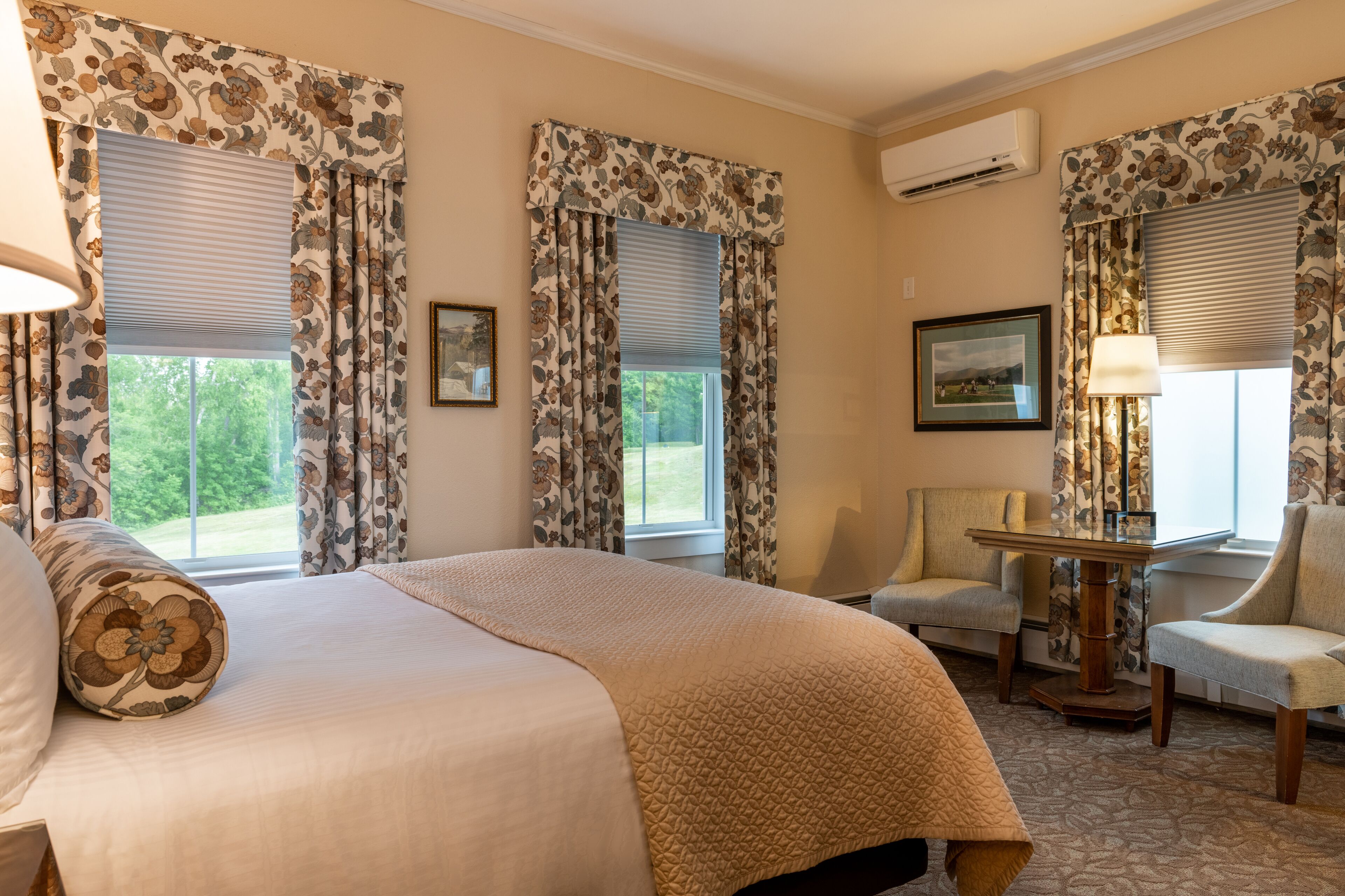 A hotel room with a large bed, two armchairs, a small table, and floral curtains.