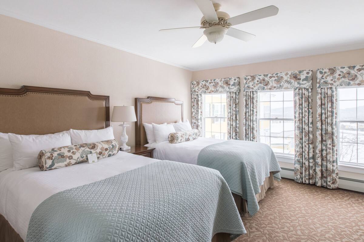 A hotel room with two beds, a ceiling fan, floral curtains, and a view of the city.