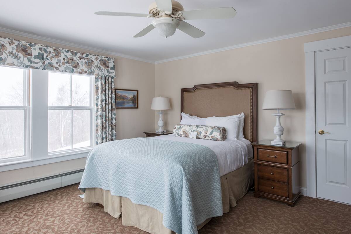 A bedroom with a bed, two bedside tables, a ceiling fan, a window with curtains, and a door.