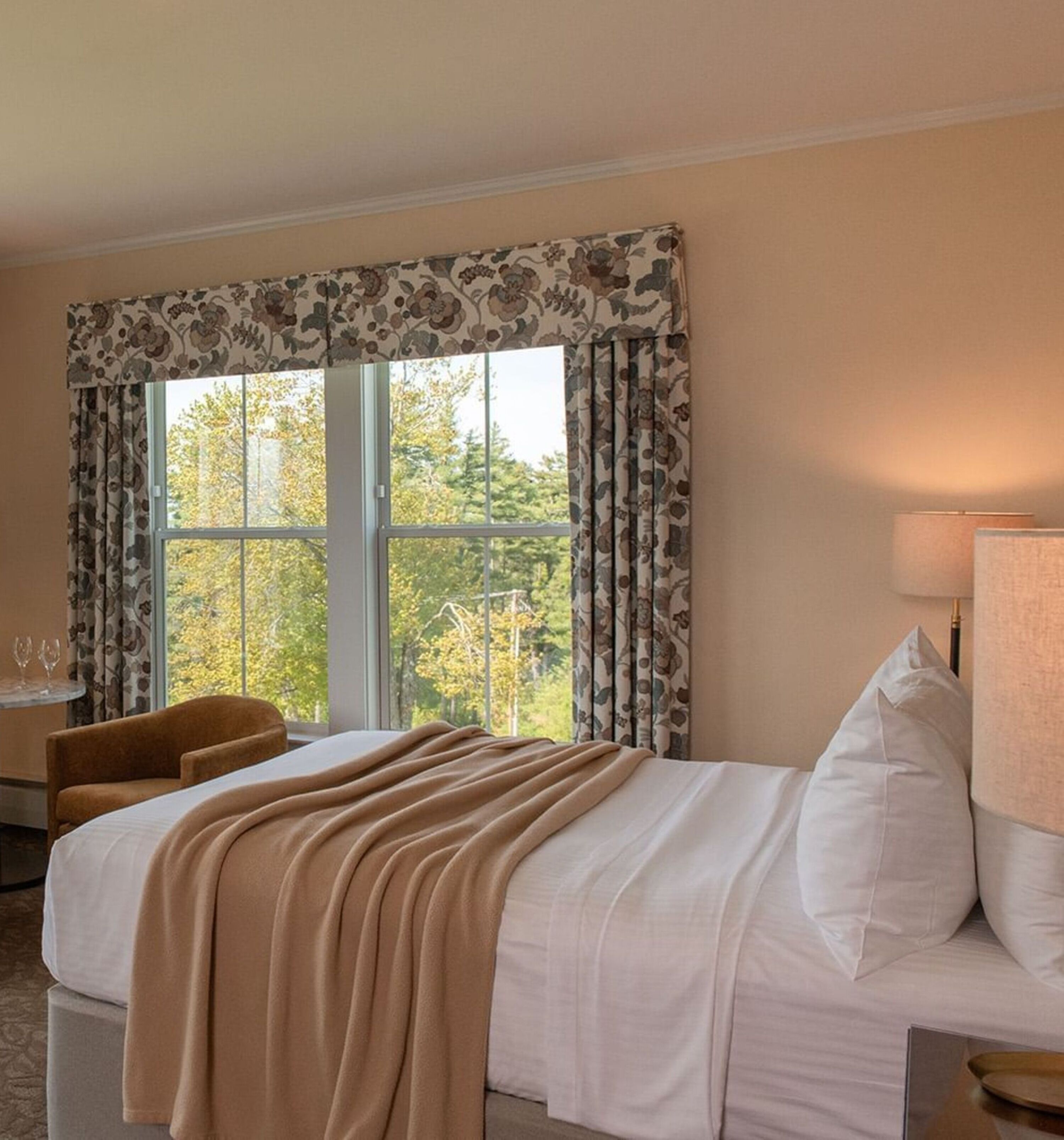 A hotel room with a bed, a chair, a table with glasses, and a lamp.