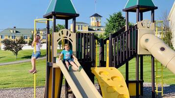 A playground with a slide and climbing structures, surrounded by a well-maintained lawn and residential buildings in the background.