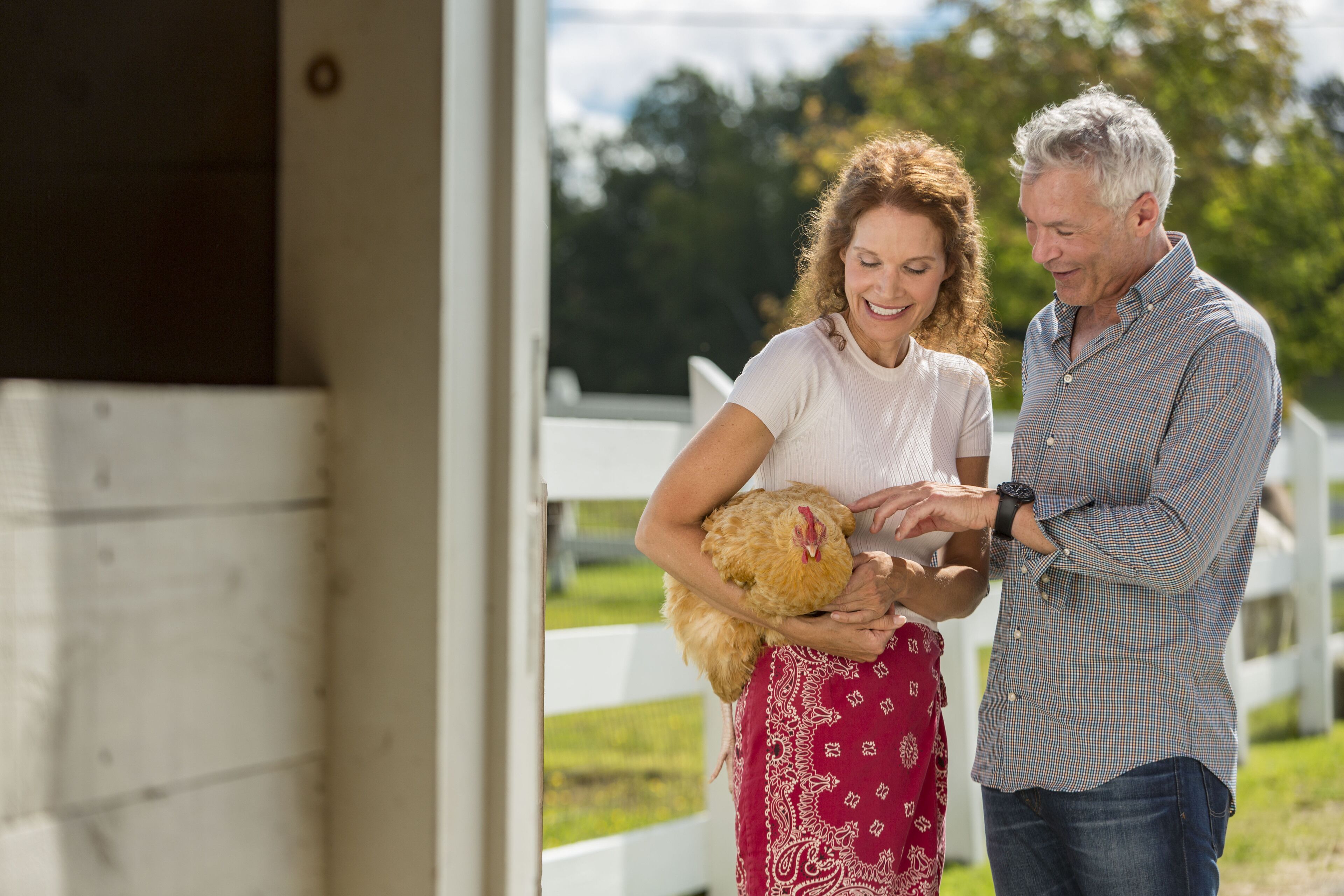 A couple holding a chicken near a white fence with a grassy area and trees in the background.
