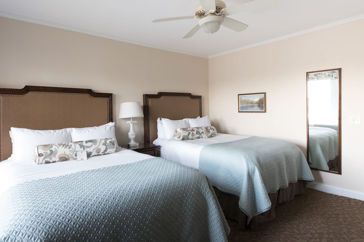 A hotel room with two beds, a ceiling fan, a bedside lamp, a framed picture on the wall, and a mirror.