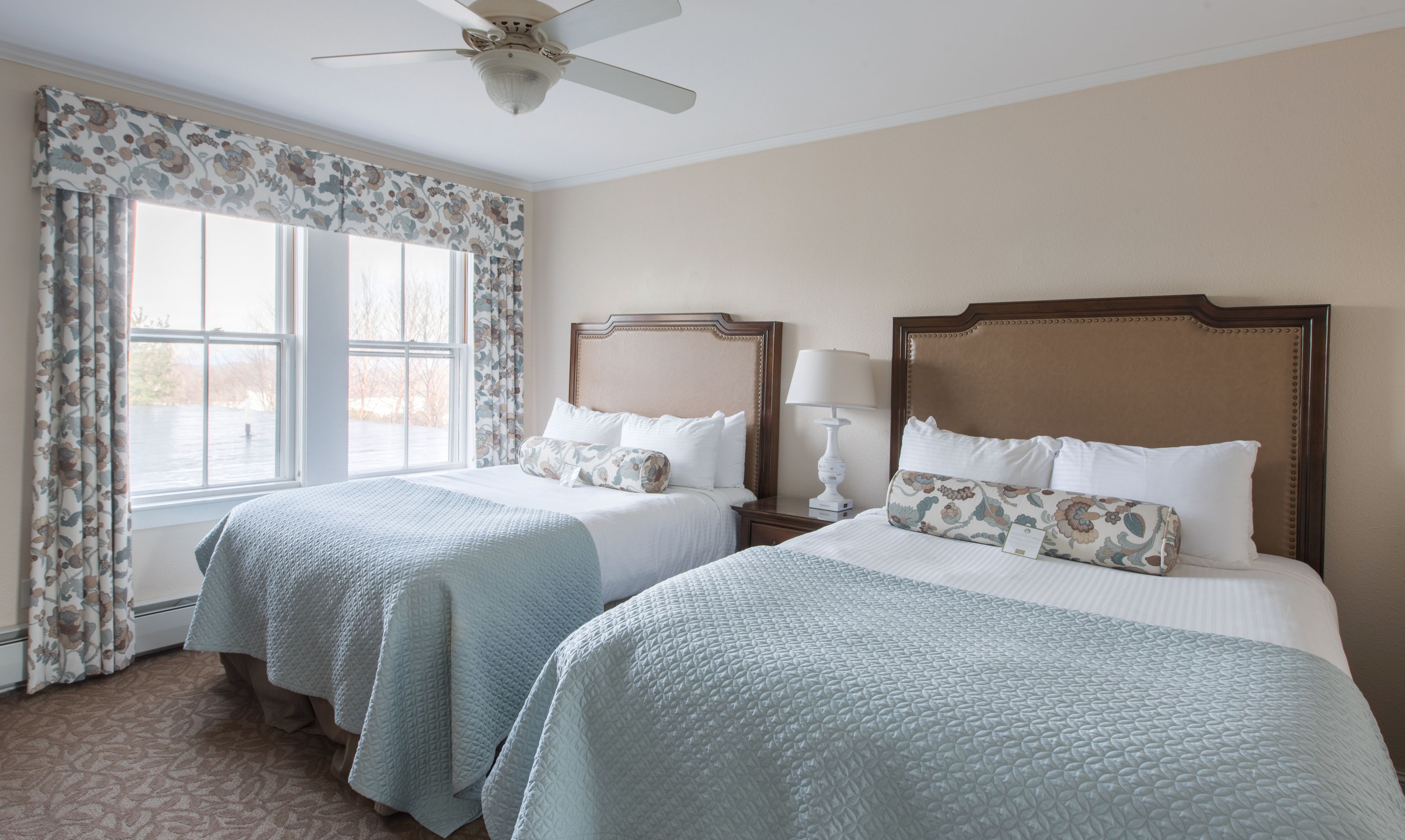 Une chambre d’hôtel avec deux lits «queen size», un ventilateur de plafond, une grande fenêtre avec des rideaux à motifs floraux et une vue sur un plan d’eau.