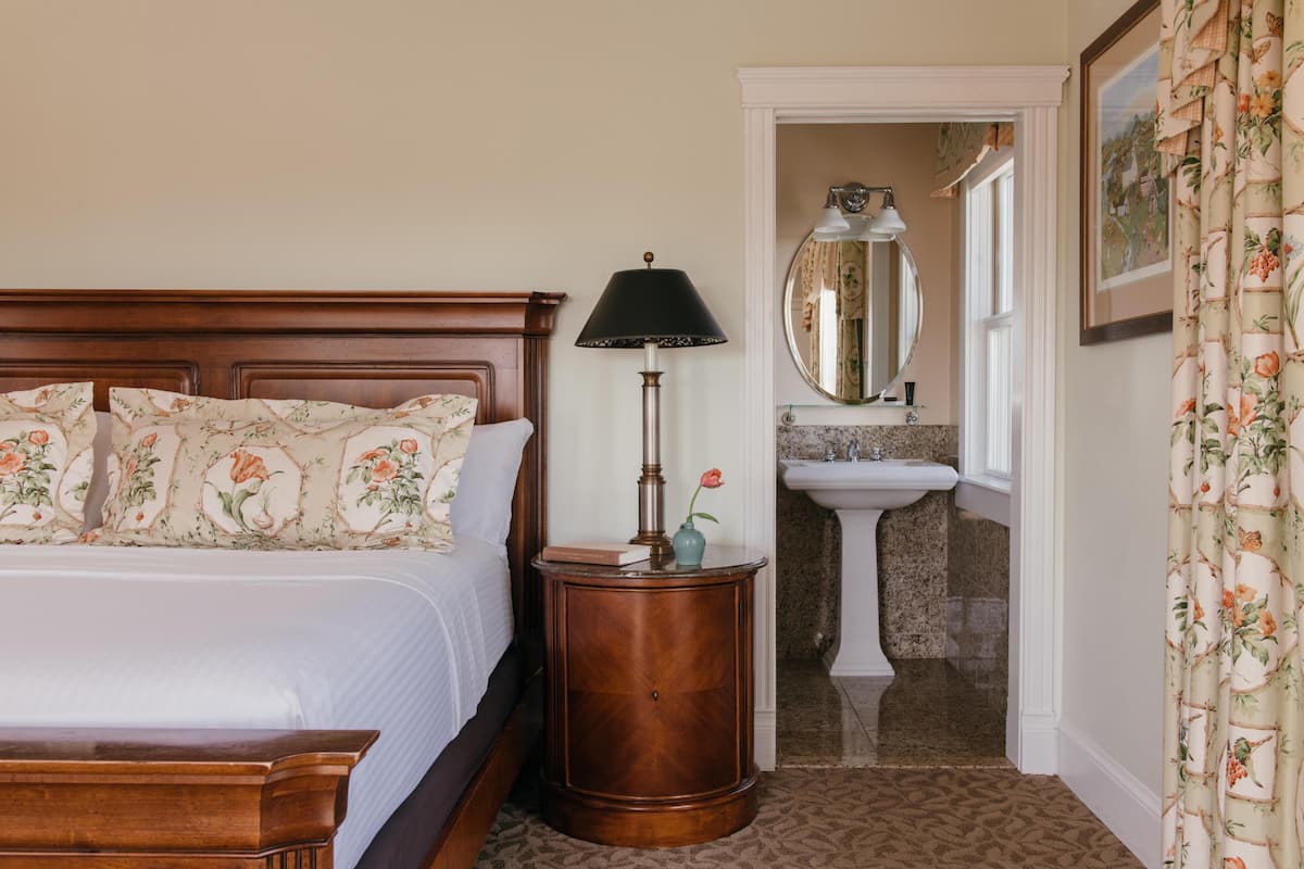 A bedroom with a wooden headboard, a bedside table with a lamp, a mirror, and a bathroom with a sink and mirror.