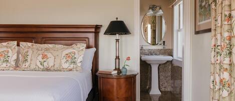 A bedroom with a wooden headboard, a bedside table with a lamp, a mirror, and a bathroom with a sink and mirror.