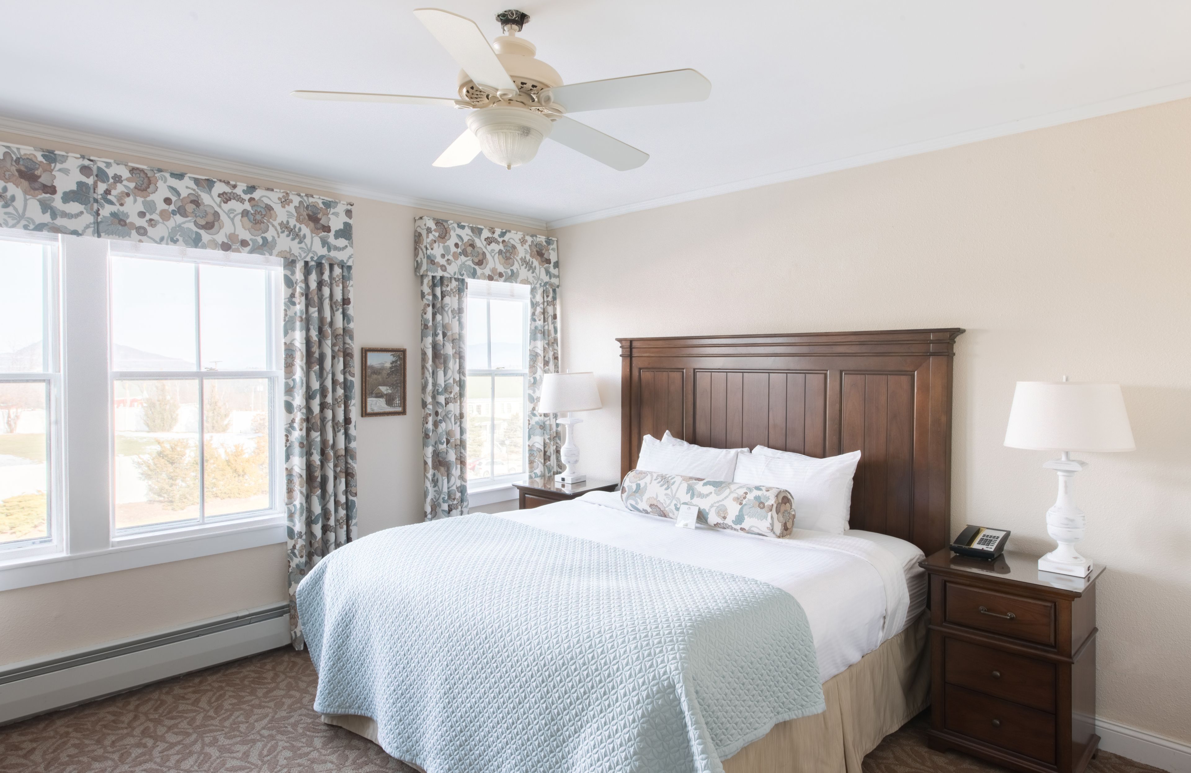 A bedroom with a large bed, two bedside tables with lamps, a ceiling fan, and a window with curtains.