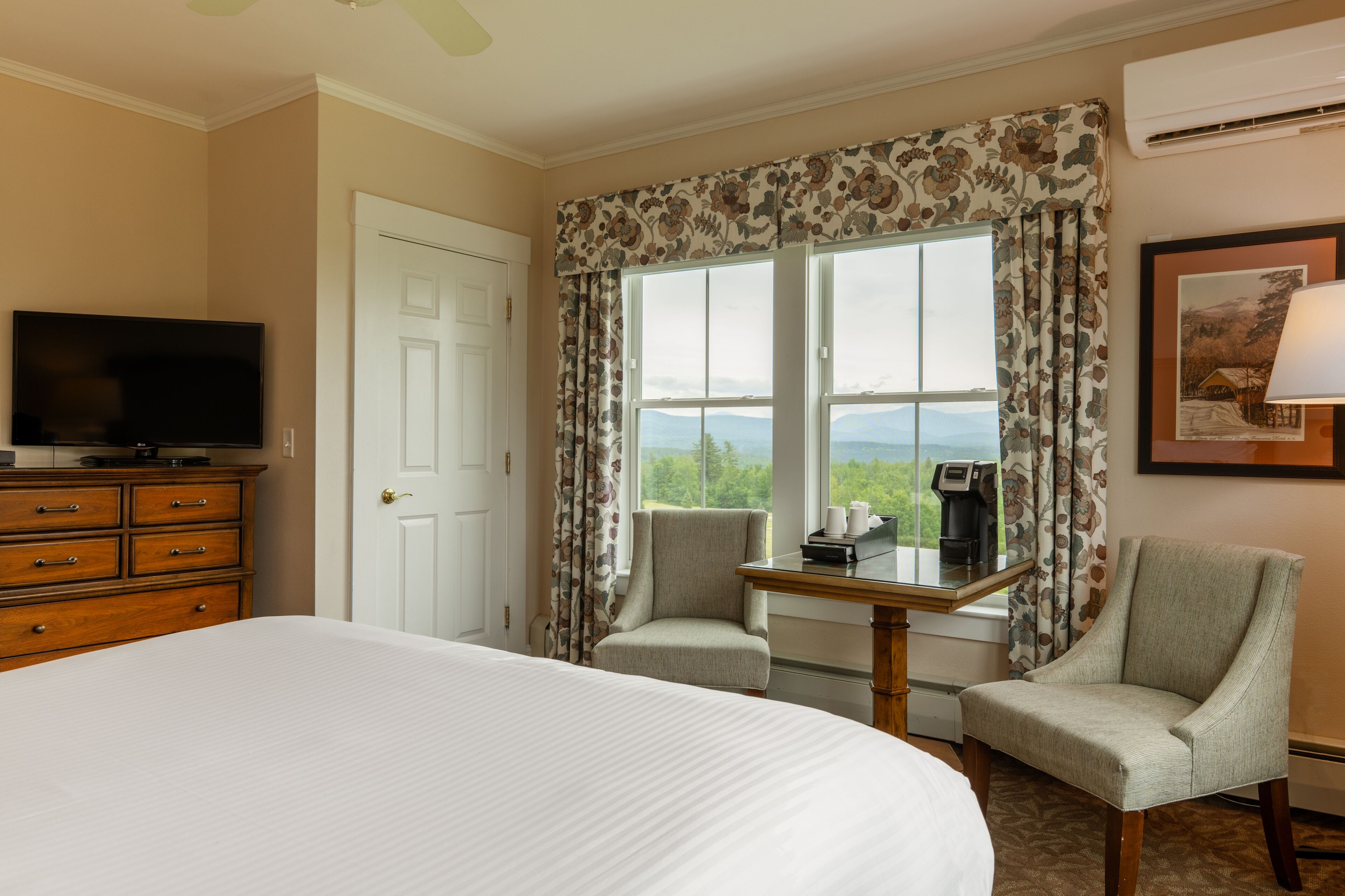 A hotel room with a large bed, a TV on a dresser, two armchairs, a small table with a coffee maker, and a window with curtains.