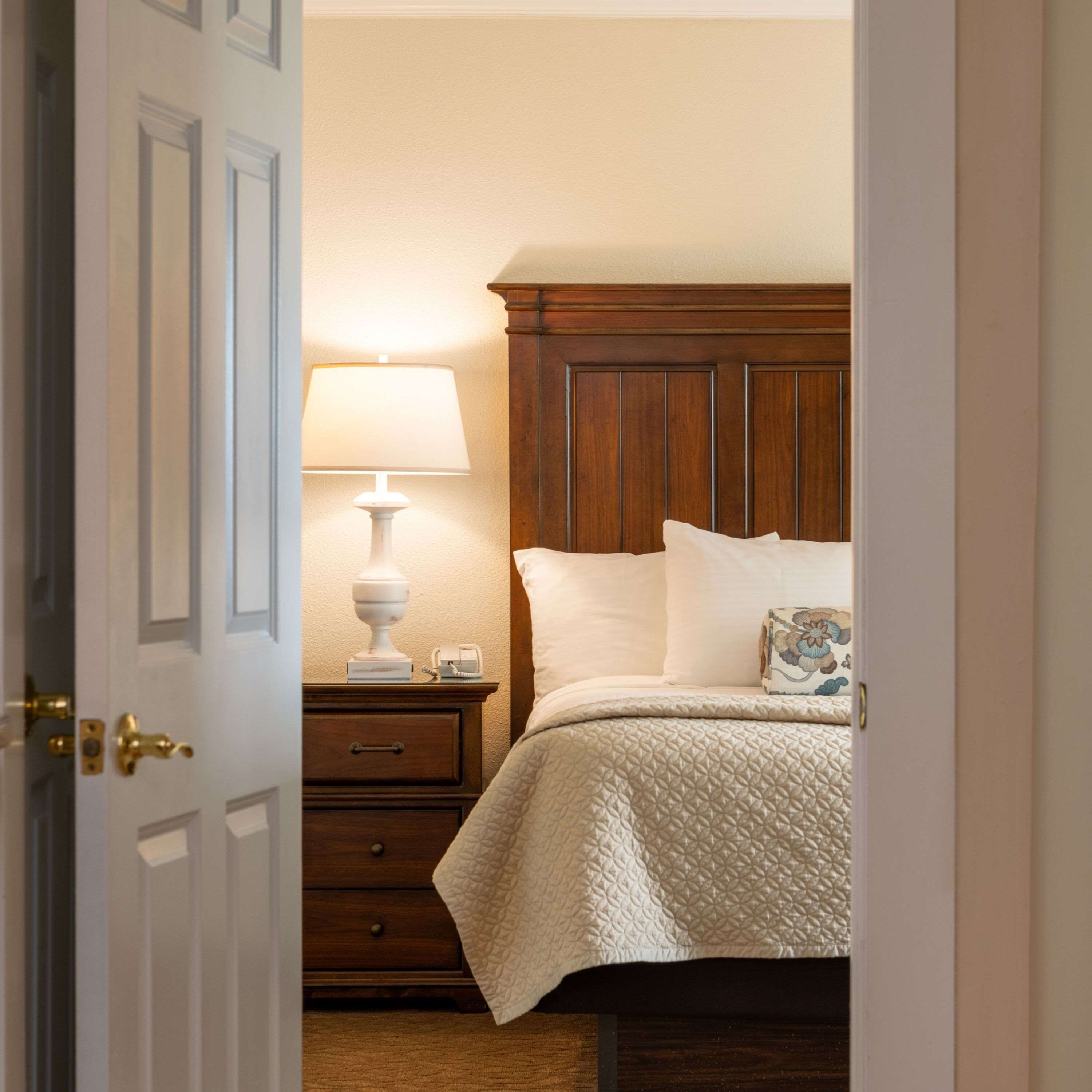 A bedroom with a wooden headboard, a bedside table with a lamp, and a bed with pillows and a quilted blanket.