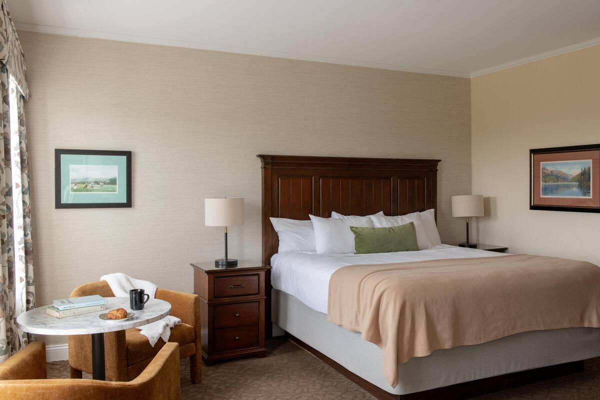 A hotel room with a bed, bedside tables, a small round table with a chair, and two framed pictures on the walls.