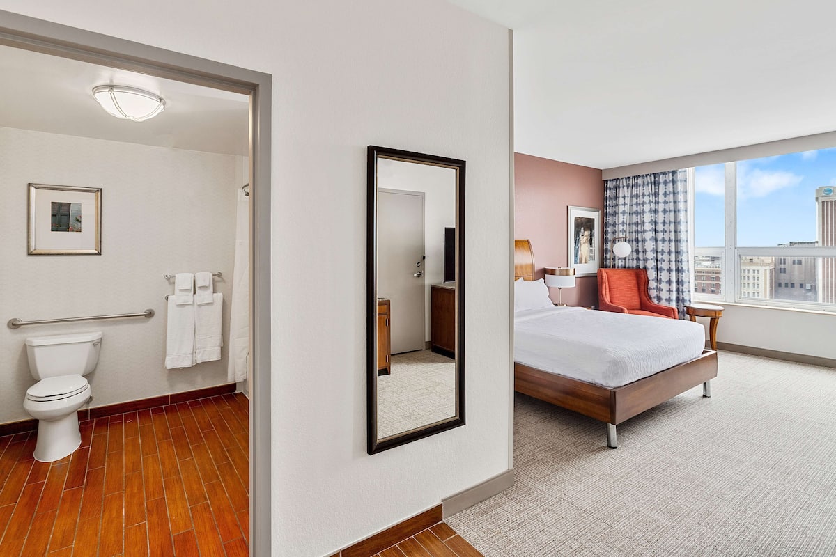 Habitación de hotel con baño, una cama grande, una mesita de noche, una ventana con vista a la ciudad y un espejo.