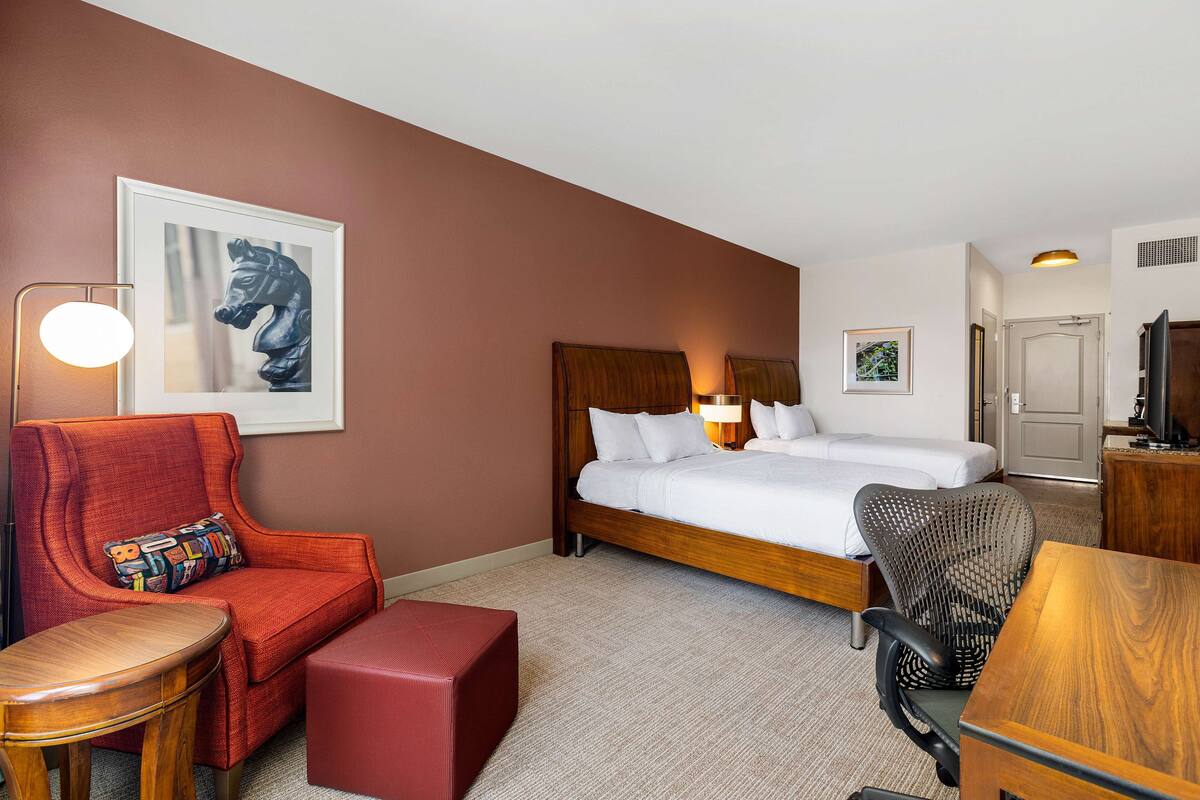 Habitación de hotel con una cama, un sillón rojo, una mesa de madera, un taburete pequeño y una lámpara de pared.