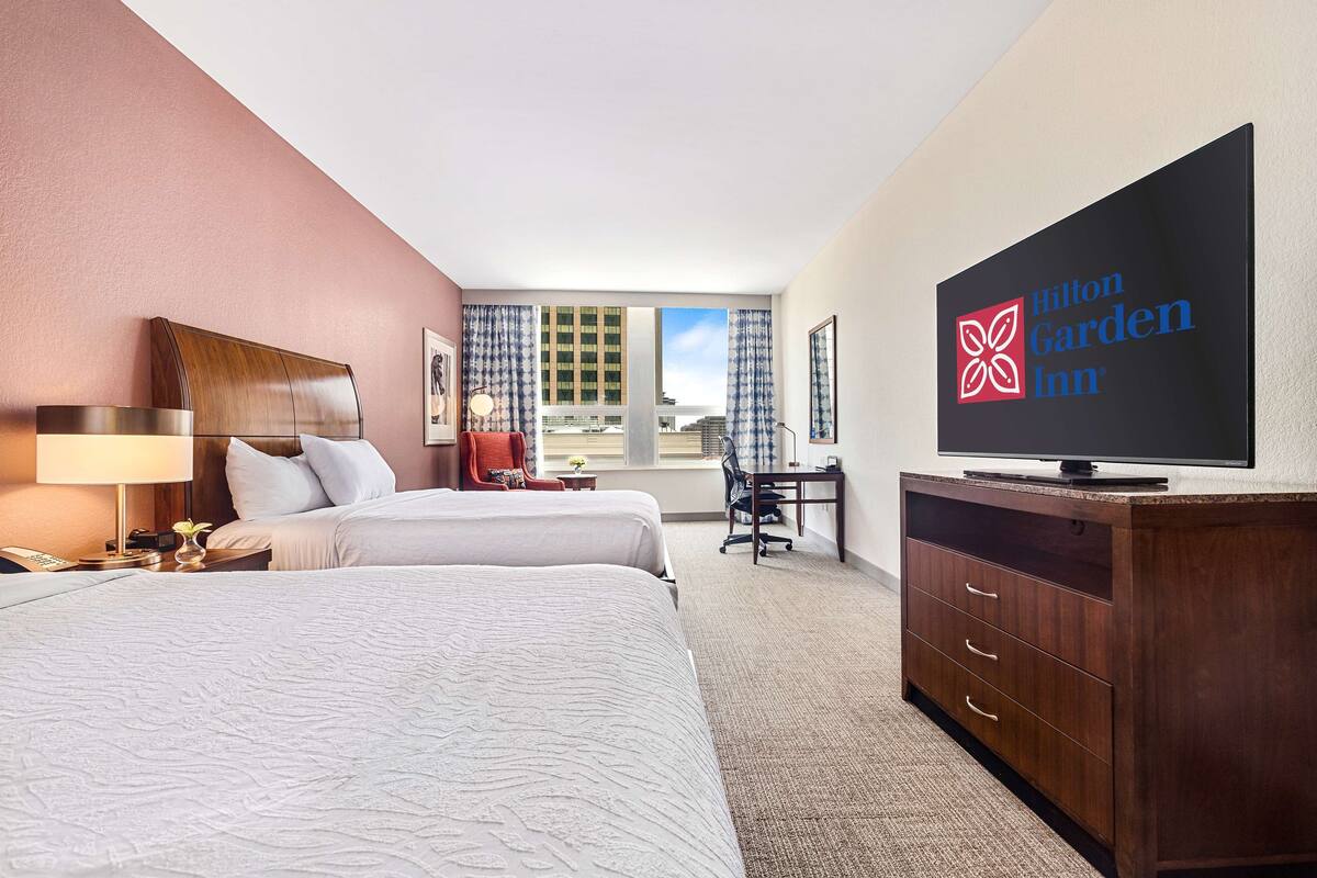 Habitación de hotel con dos camas, un escritorio, una silla, un televisor y vistas a la ciudad.