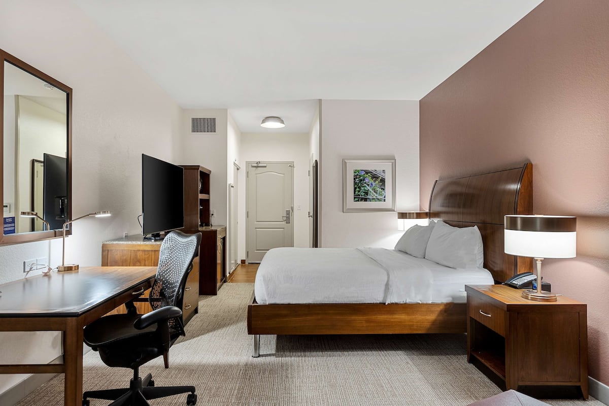 Habitación de hotel con cama, escritorio y silla, televisor, mesita de noche con lámpara, y puerta que da a otra habitación.