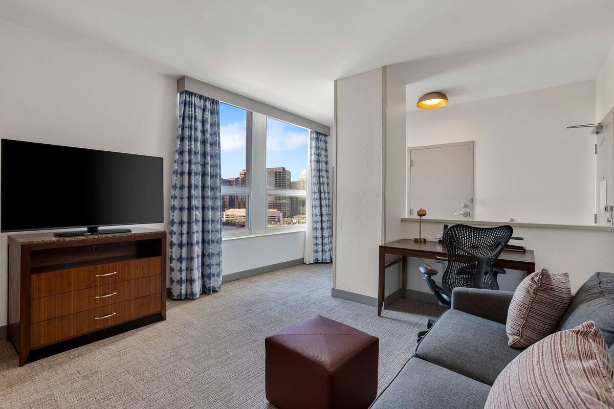 Habitación de hotel con televisión de pantalla plana, escritorio con silla, sofá y ventana grande con cortinas.