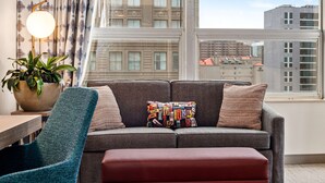 Ein modernes Wohnzimmer mit einem grauen Sofa, einem farbenfrohen Kissen, einem blauen Sessel und einem großen Fenster mit Blick auf die Stadt.